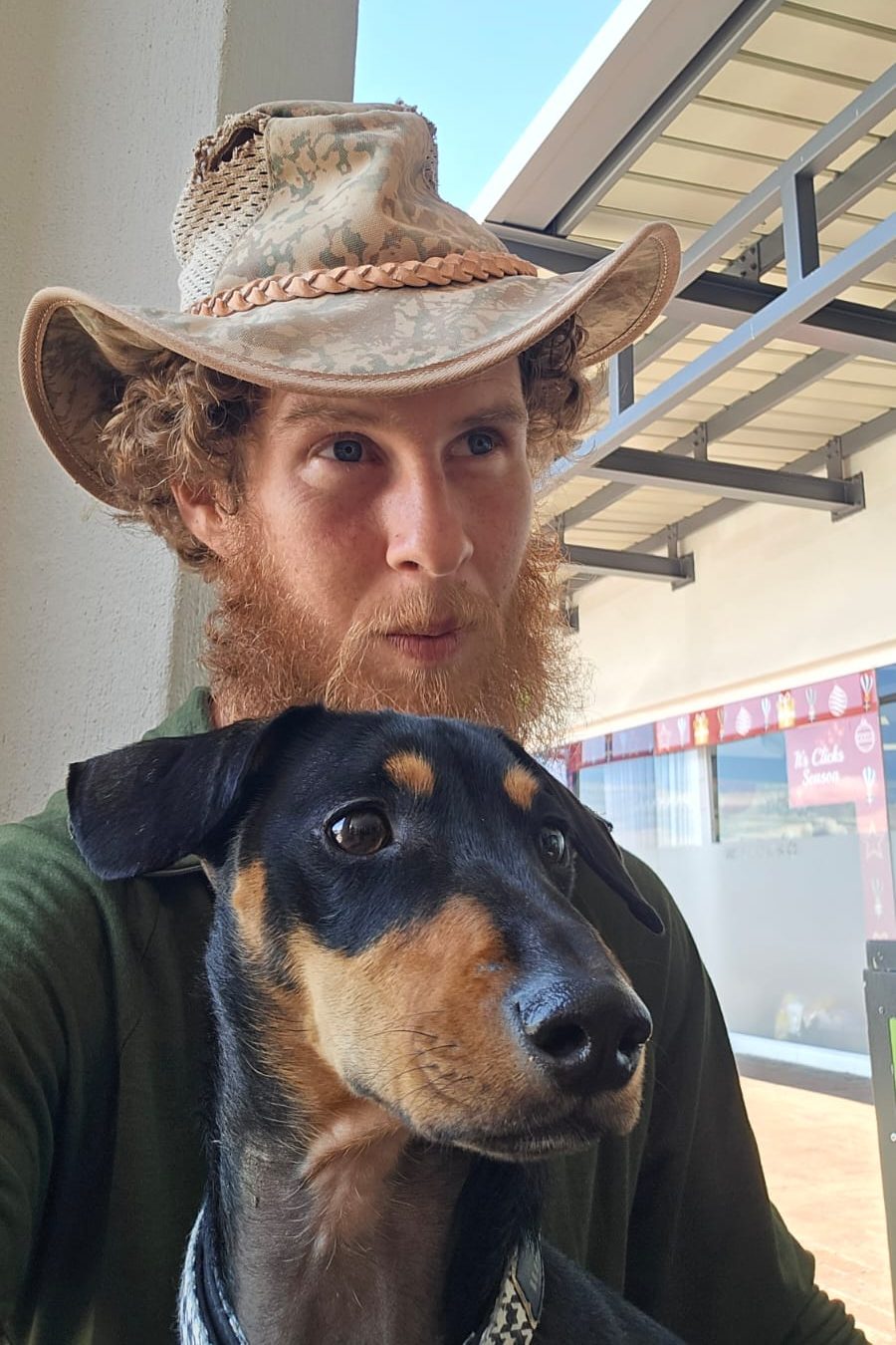 A man with curly hair and a beard wearing a wide-brimmed hat, sitting next to a black and tan dog with a curious expression.